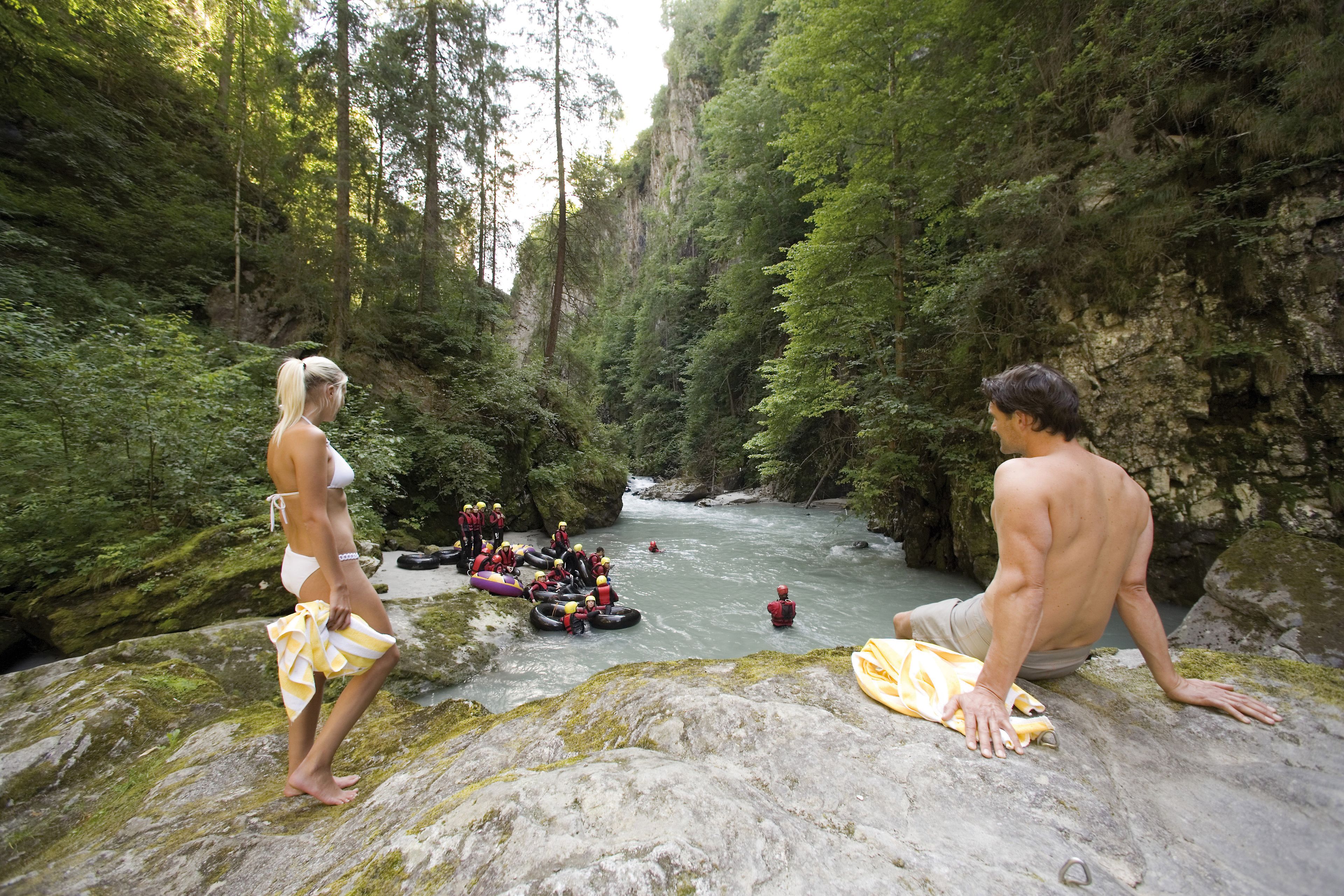 A group of people on a rocky riverbank, some in rafts and others on the shore, surrounded by dense forest.