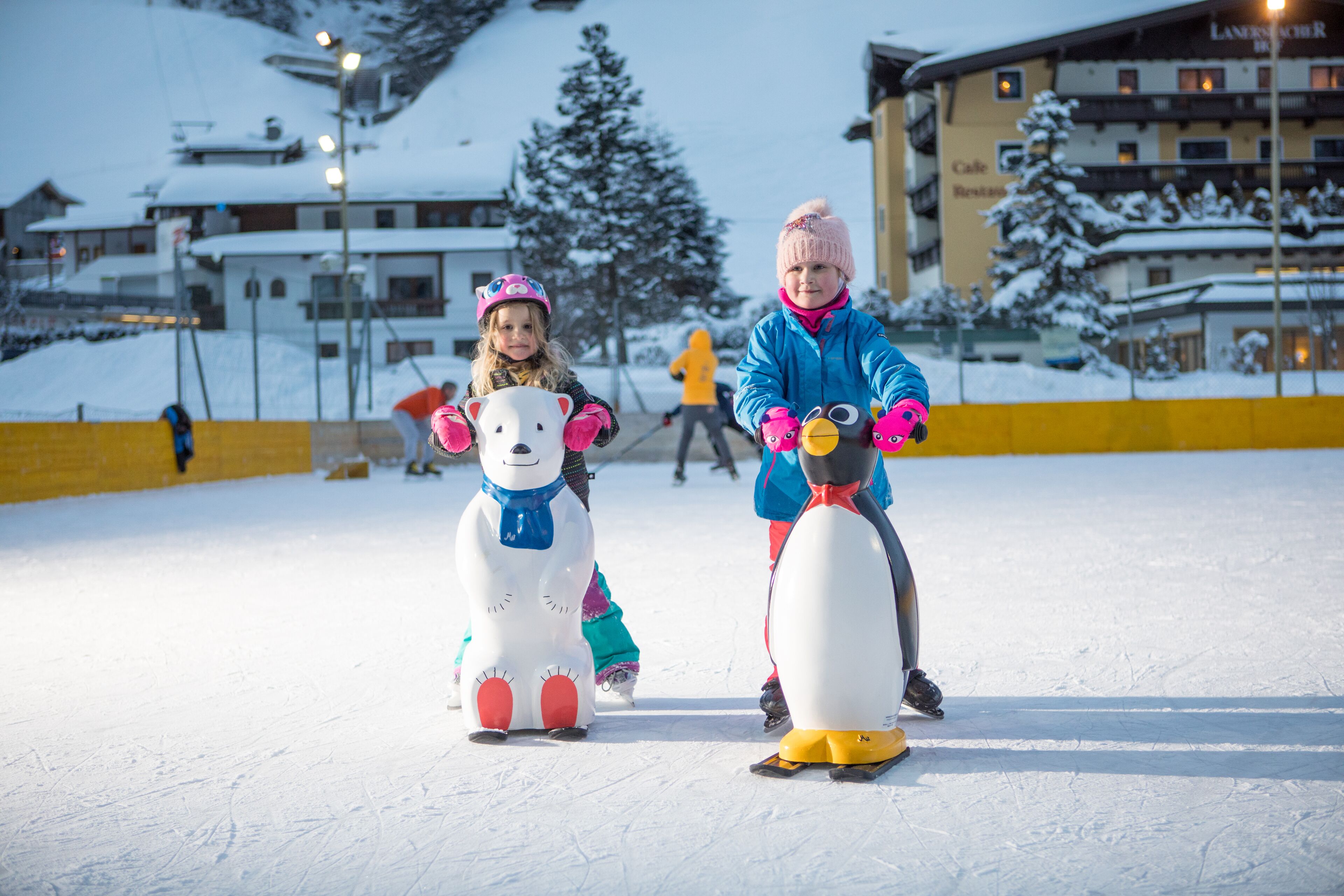 Two children on snow-themed ice skates in a snowy outdoor area with buildings in the background.