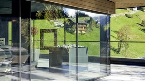 A modern interior with large glass windows offering a view of a green hillside and traditional buildings. - Hotel Tirolerhof (Tux)