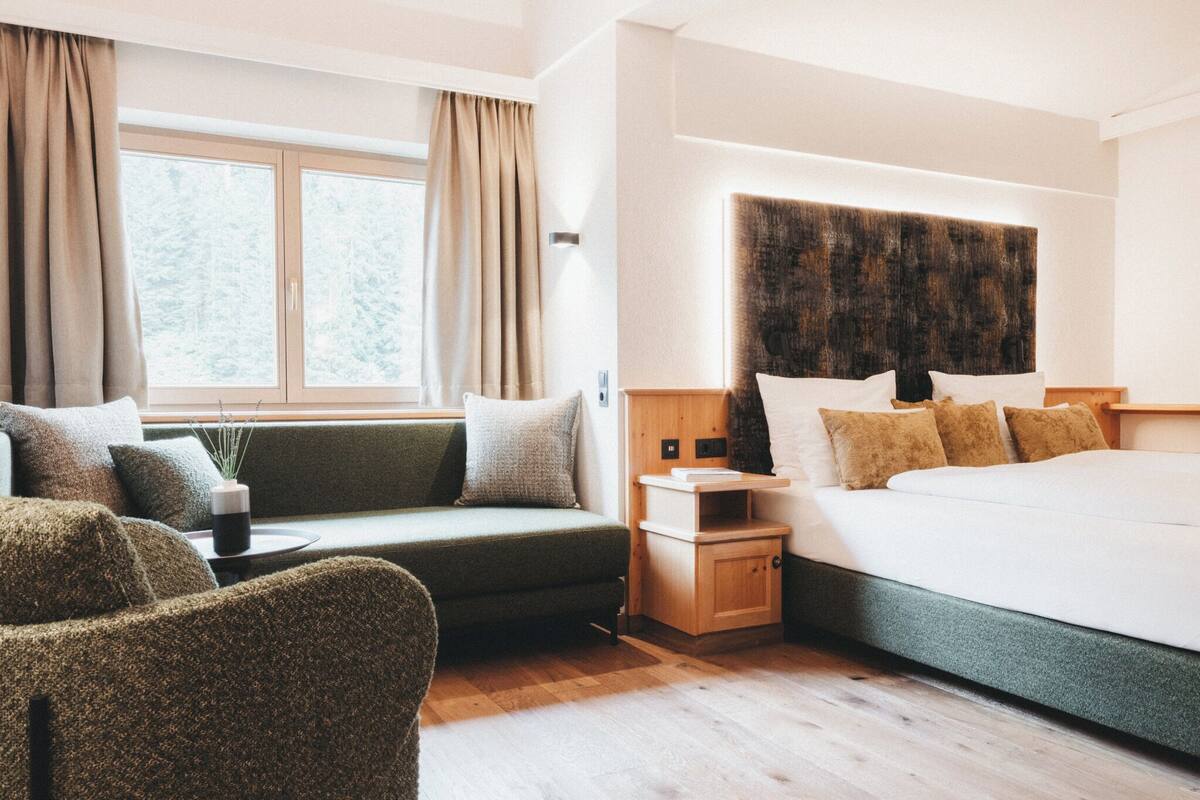 Una habitación de hotel moderna con una cama grande, un sofá, una mesita y una ventana con cortinas.
