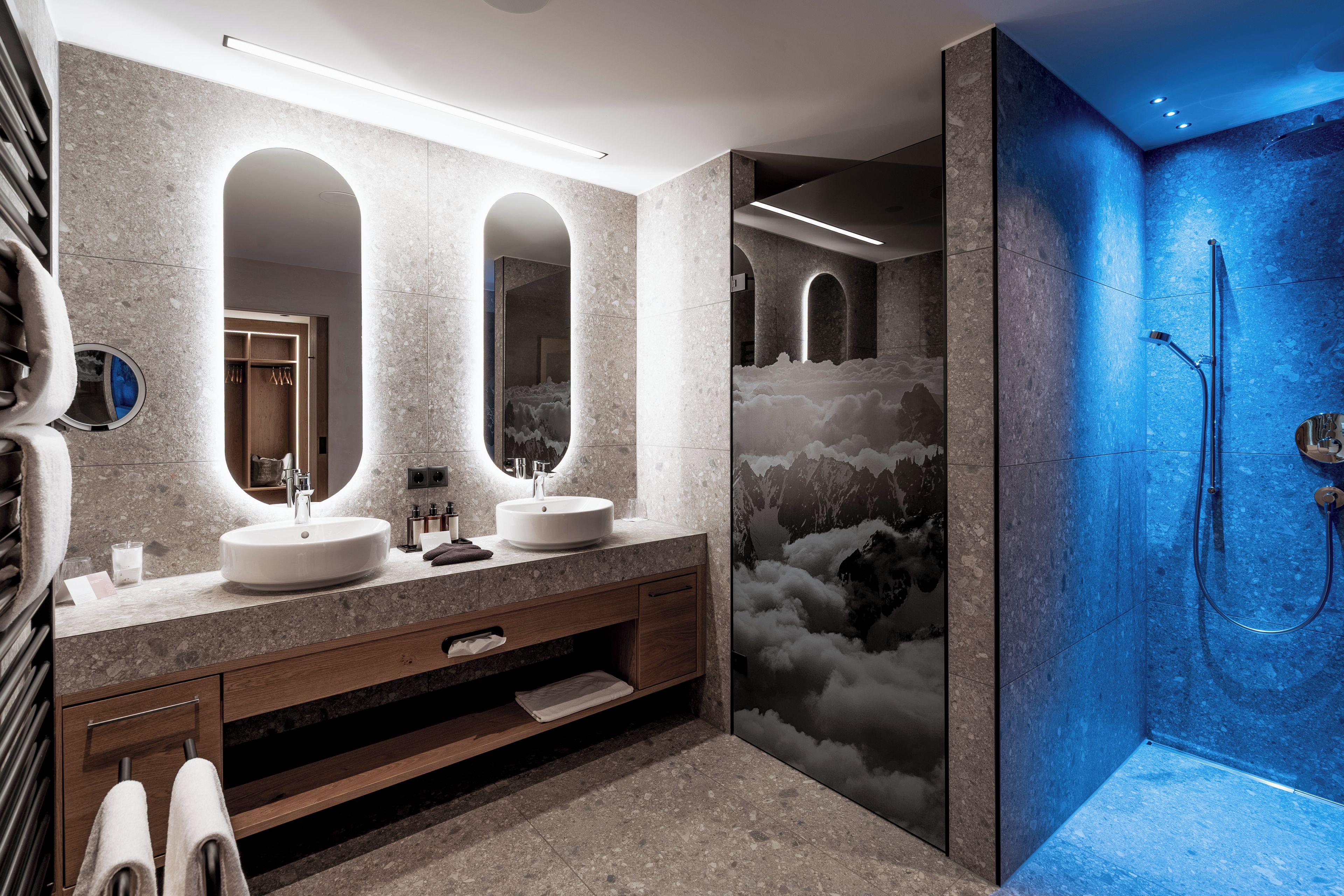 A modern bathroom with two sinks, a vanity, a mirror, and a shower with a blue-tinted glass door.