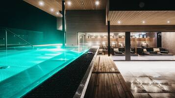 A modern indoor swimming pool with a wooden deck and lounge area.