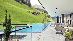 A modern outdoor pool area with a glass balustrade, lounge seating, and a view of a hillside with houses. - Hotel Tirolerhof (Tux)