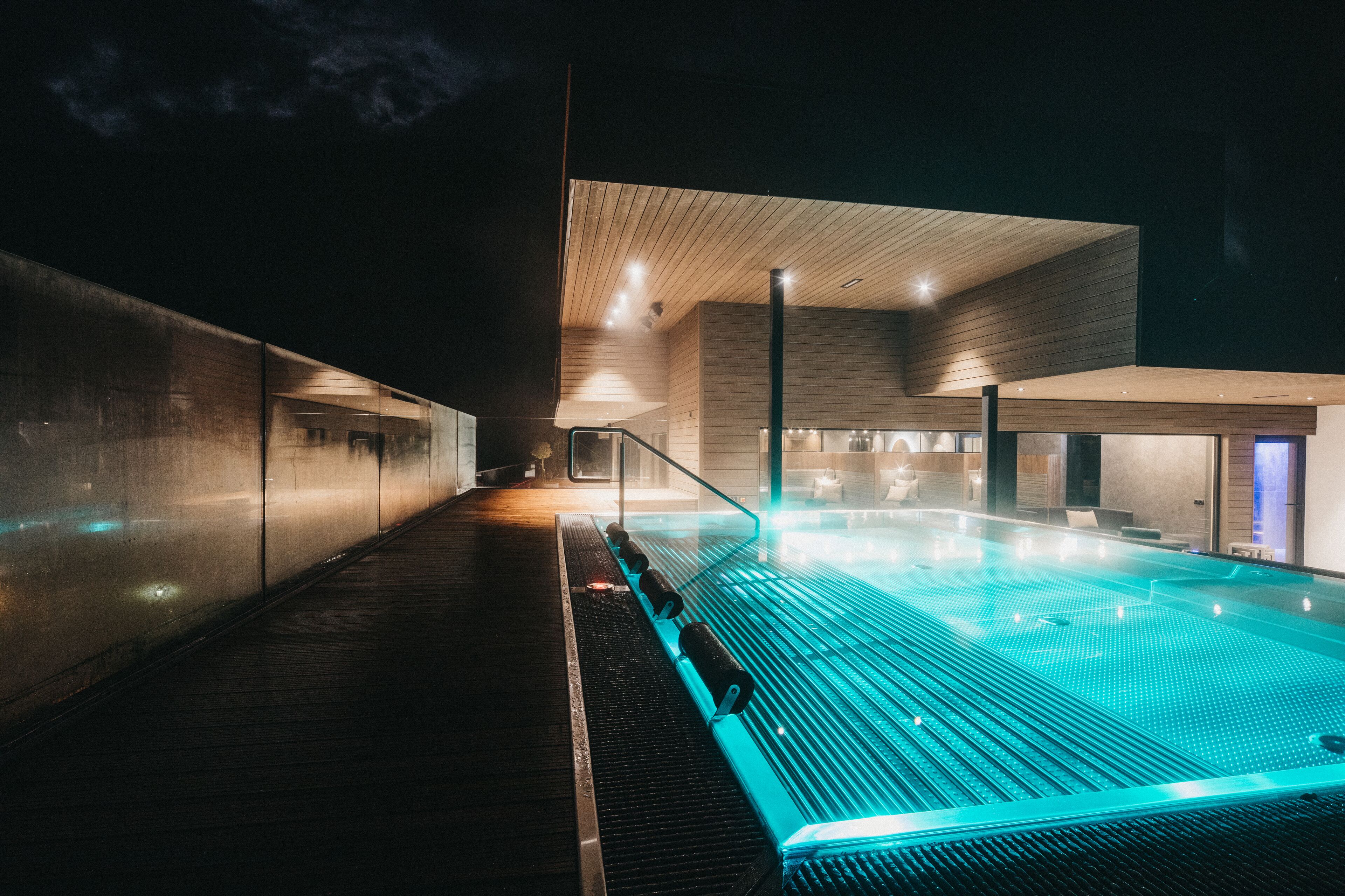Una piscina interior moderna con plataforma de madera y paredes de vidrio, iluminada con luces azules.