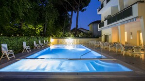 A swimming pool with lounge chairs and tables, surrounded by trees and a building. - Hotel Athena (Cervia)