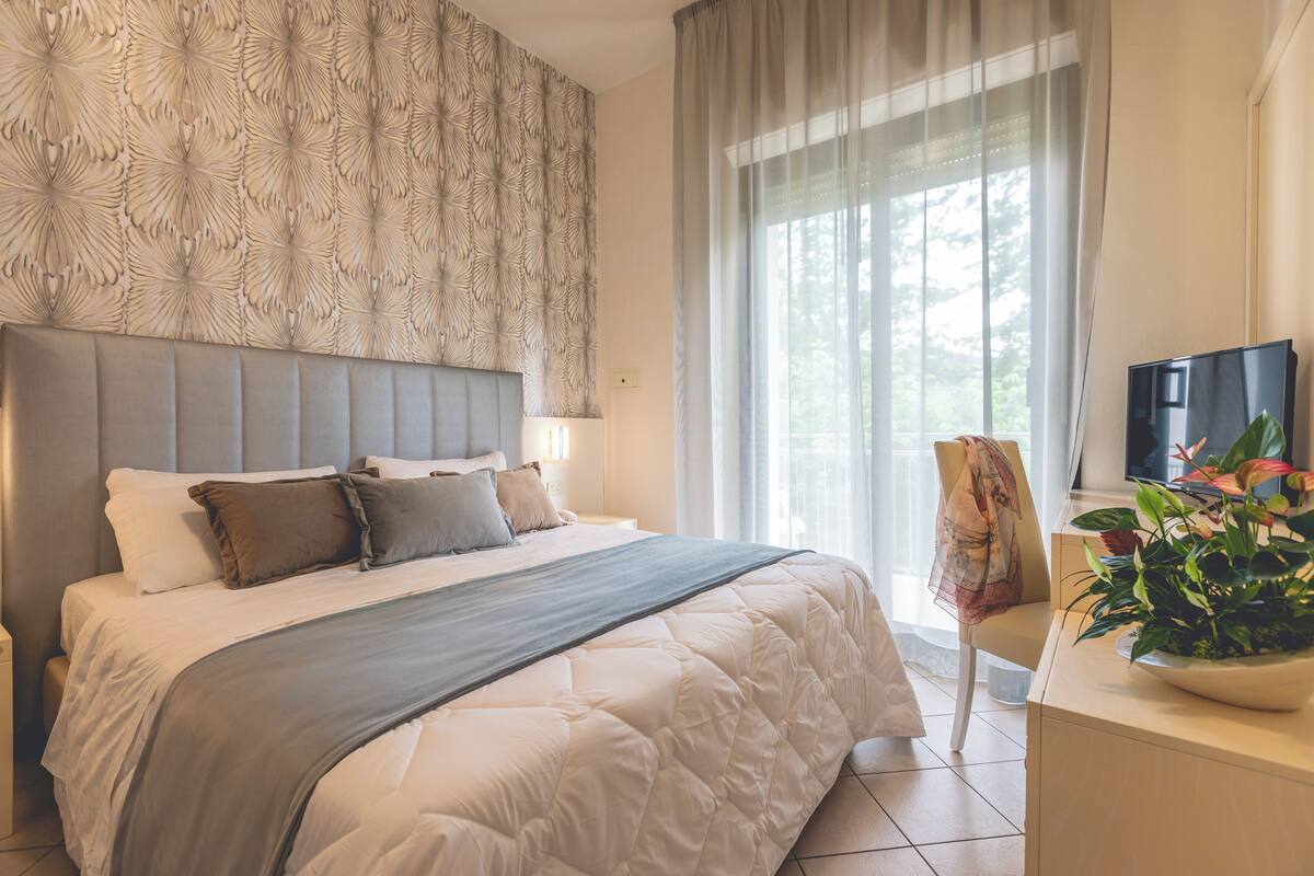A bedroom with a large bed, a headboard, a bedside table, a television, and a window with curtains.