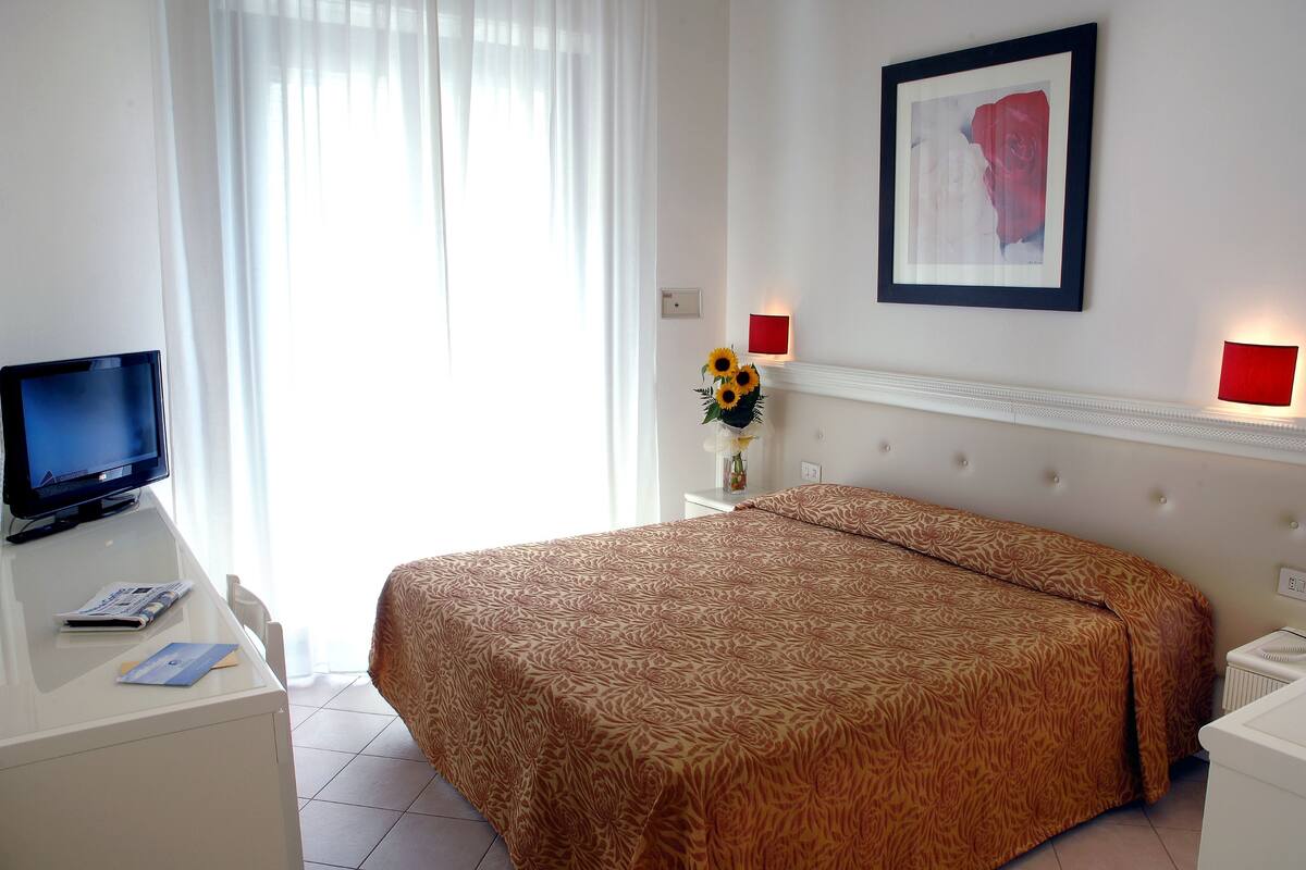 A hotel room with a bed, a desk with a television, and a vase with sunflowers.