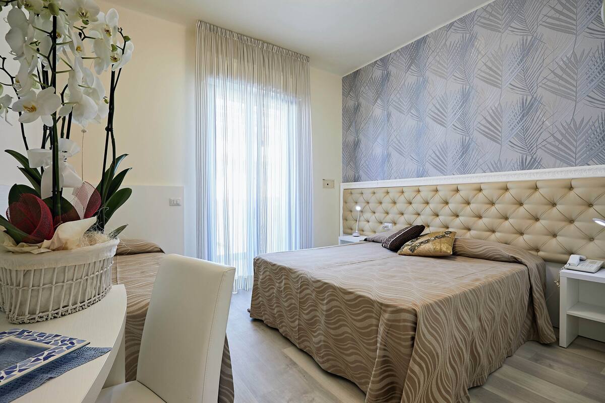 A hotel room with a tufted headboard, a bed with a patterned bedspread, a bedside table with a lamp, and a large window with sheer curtains.