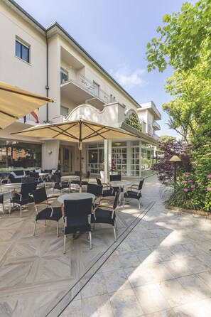 An outdoor patio with tables and chairs, a covered seating area, and a building with a balcony. - Hotel Athena (Cervia)