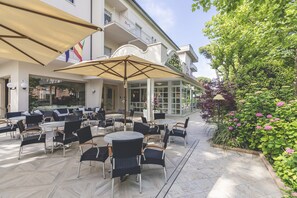 An outdoor seating area with round tables and chairs, large umbrellas, and a view of a building with balconies and a garden. - Hotel Athena (Cervia)