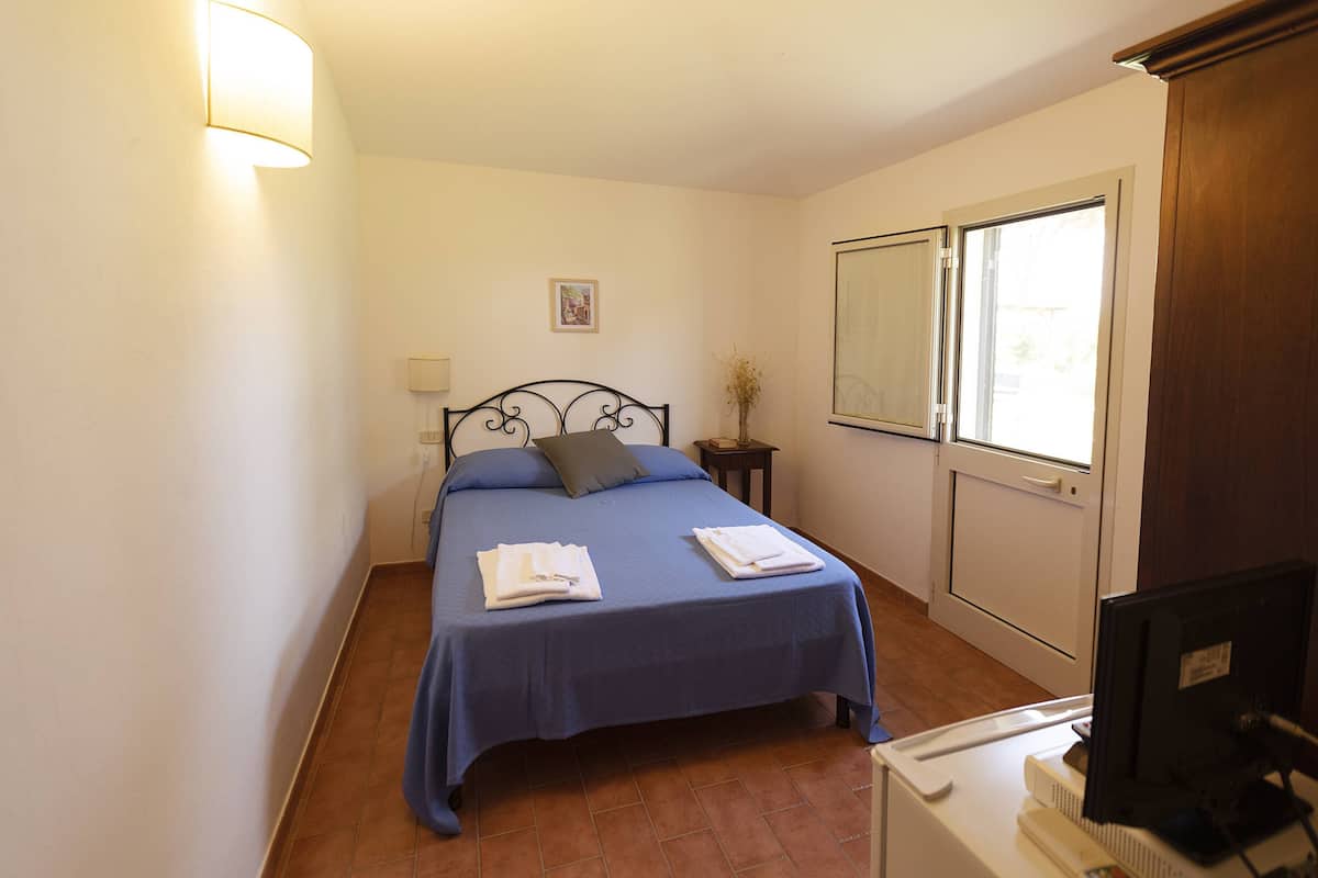 A bedroom with a bed, a bedside table, a lamp, a window, and a computer desk.