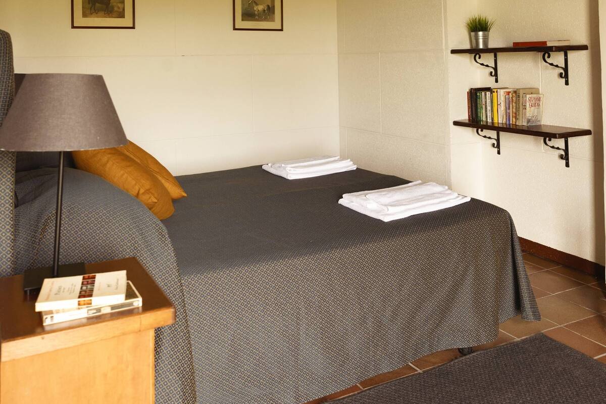 A room with a bed, a bedside table with a lamp, a shelf with books, and framed pictures on the wall.