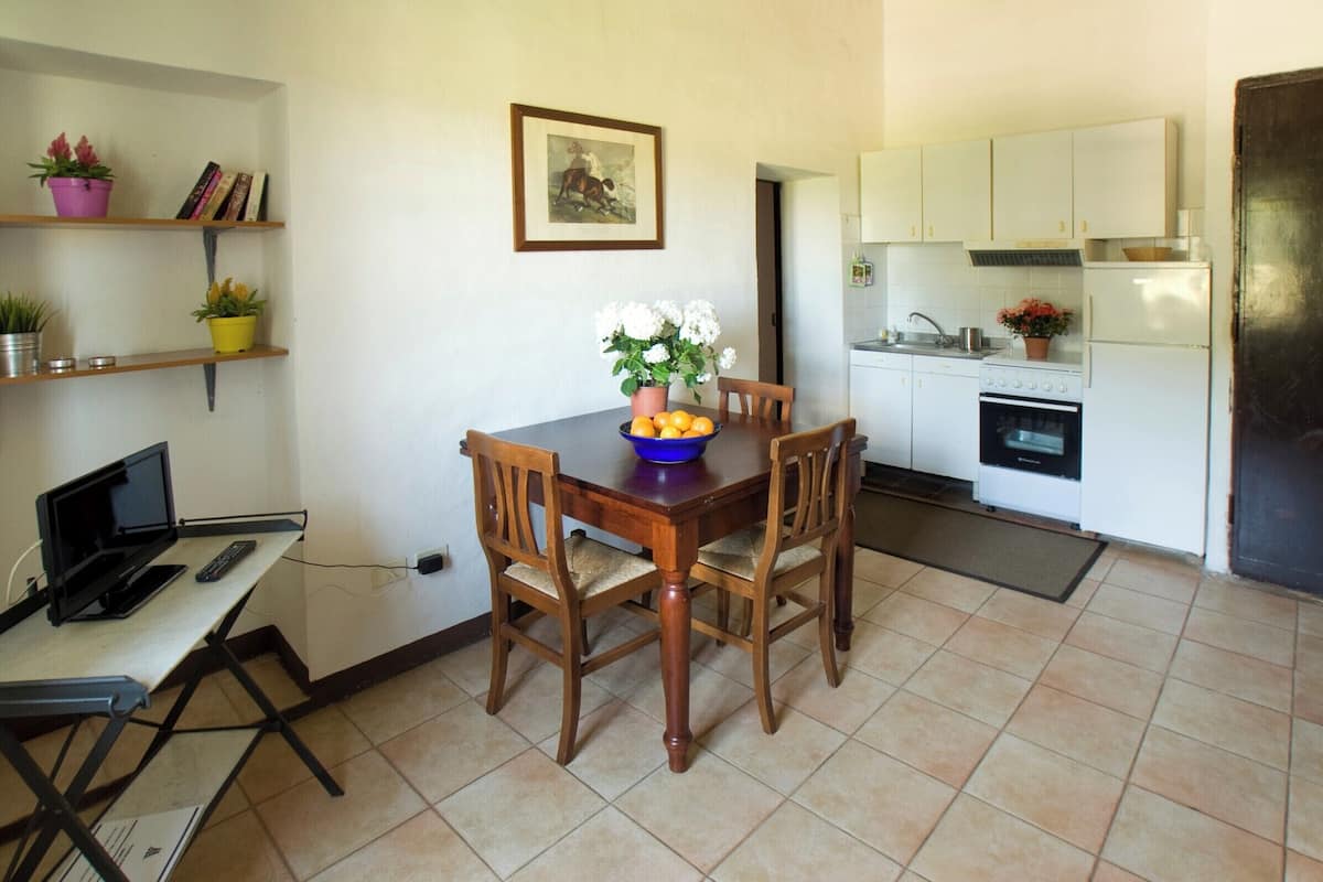 A compact kitchen with white cabinets, a stove, and a sink. There is a dining table with chairs and a bowl of fruit.
