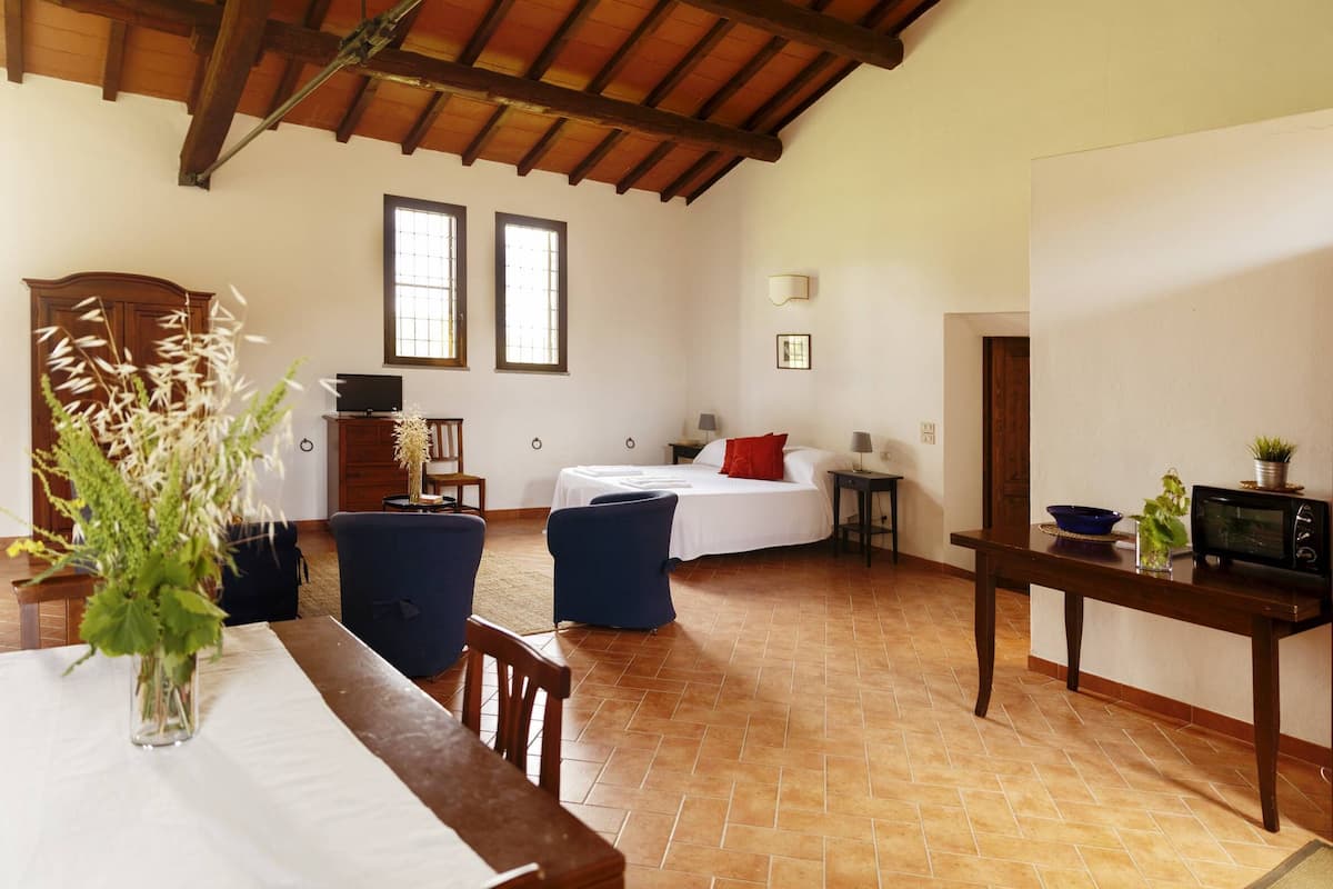 A room with a bed, a table with a vase, a chair, a TV, a lamp, and a microwave.
