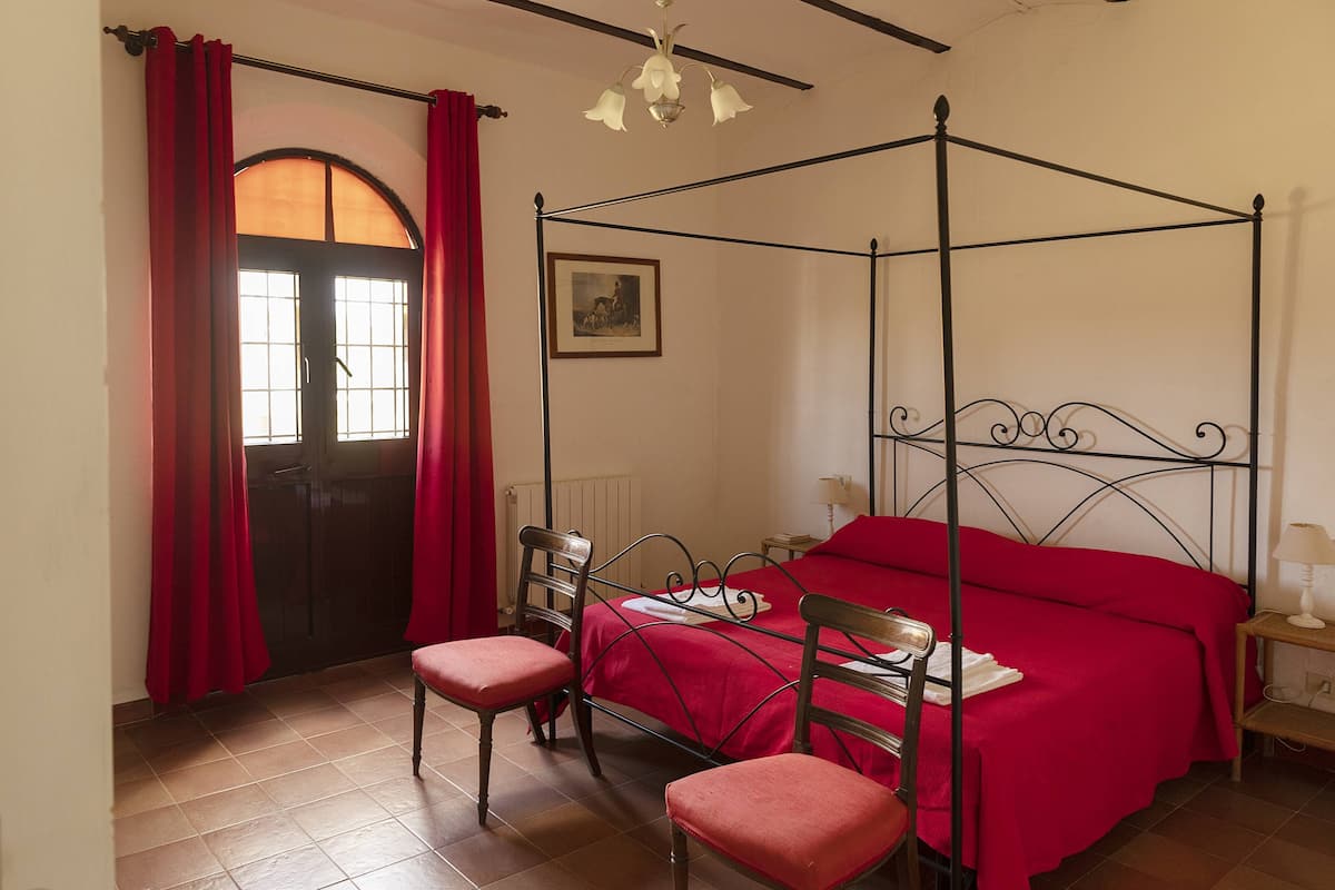 A four-poster bed with a red bedspread, two wooden chairs, a red curtain, and a framed picture on the wall.