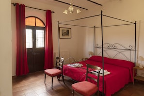 Ein Himmelbett mit roter Bettdecke, zwei Holzstühle, ein roter Vorhang und ein gerahmtes Bild an der Wand.