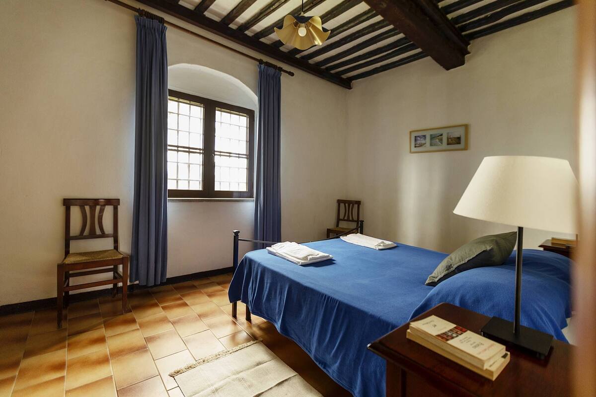 A room with a bed, a chair, a window with curtains, a lamp, and a small table with a book.