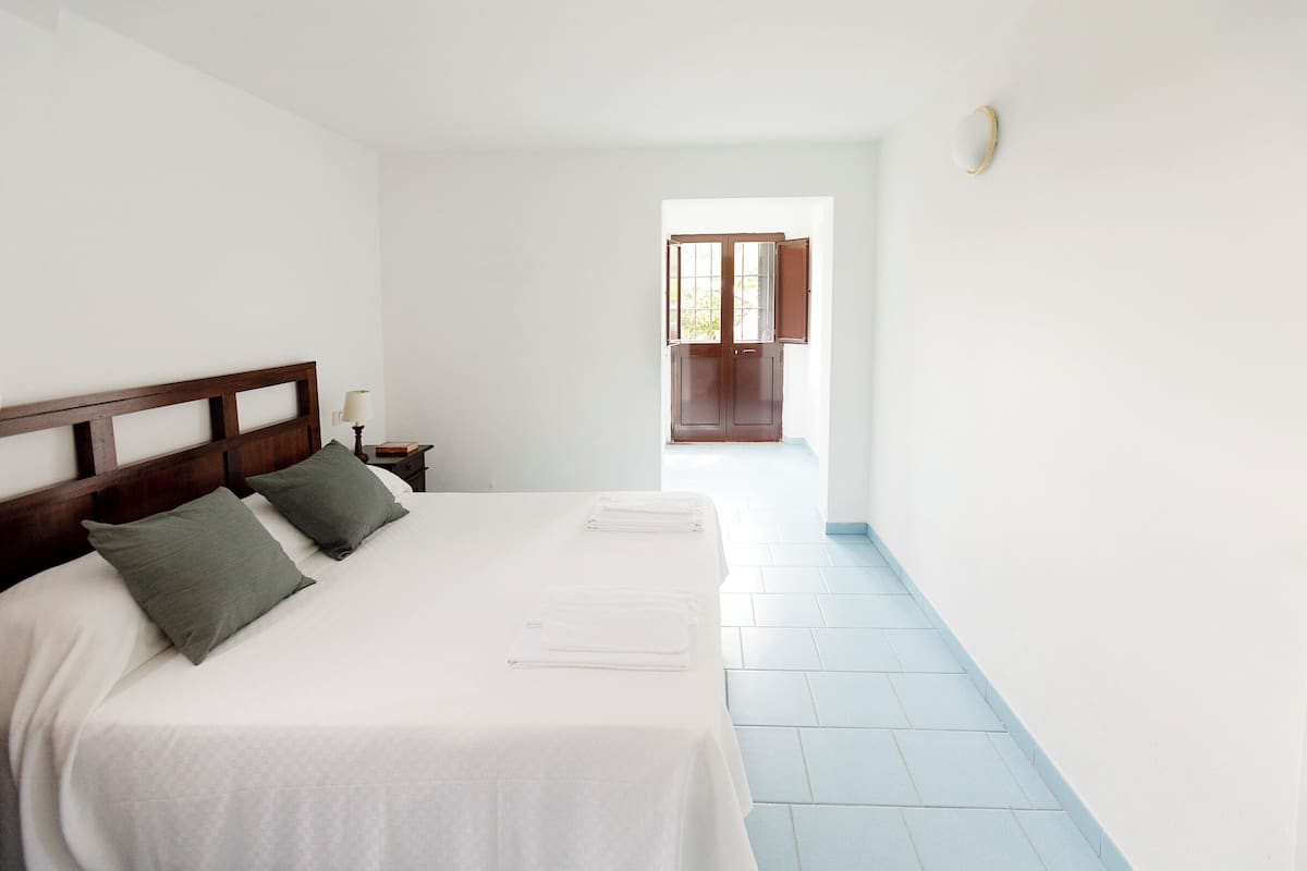 A hotel room with a large bed, two pillows, a nightstand, a window with wooden shutters, and a light fixture on the wall.