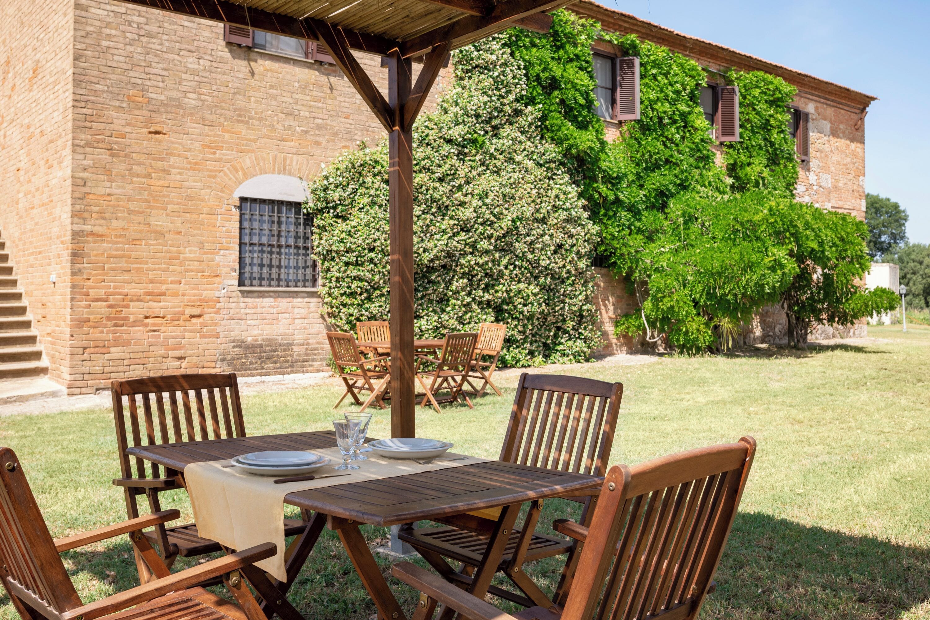An outdoor wooden table set with plates and glasses, surrounded by chairs, in front of a brick building with a covered patio area.