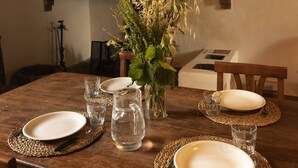 Una mesa de comedor de madera dispuesta con platos, cubiertos y vasos de agua, con un florero de plantas secas en el centro.