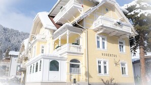 A multi-story hotel with a yellow facade, named 'HUBERTUS'. It features balconies on each floor, a central entrance, and a view of snow-covered trees in the background. - Hotel Garni Hubertus (Fulpmes)