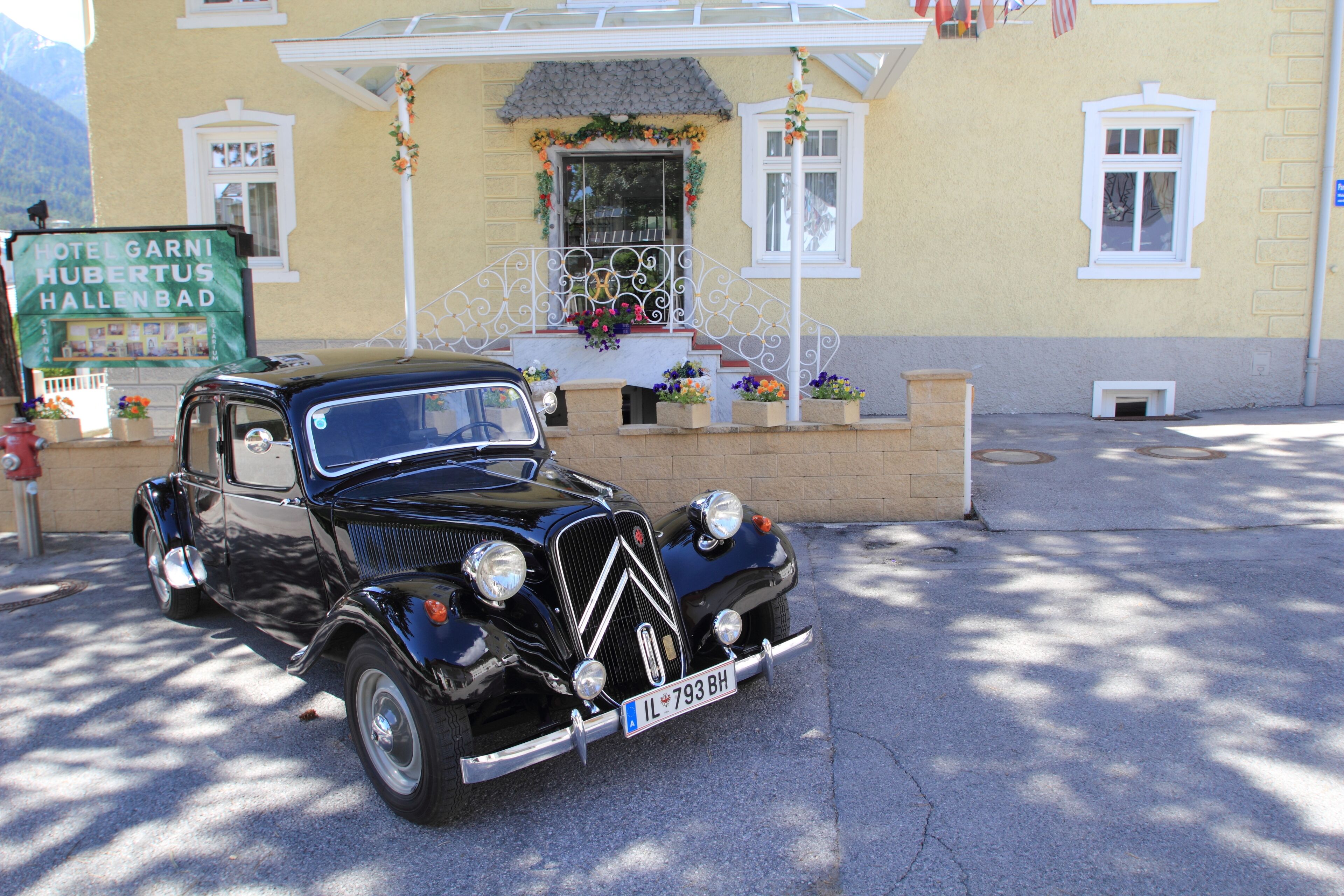 A vintage black car parked in front of a yellow building with a sign reading "Hotel Garnis Hubertus Hallenbad".