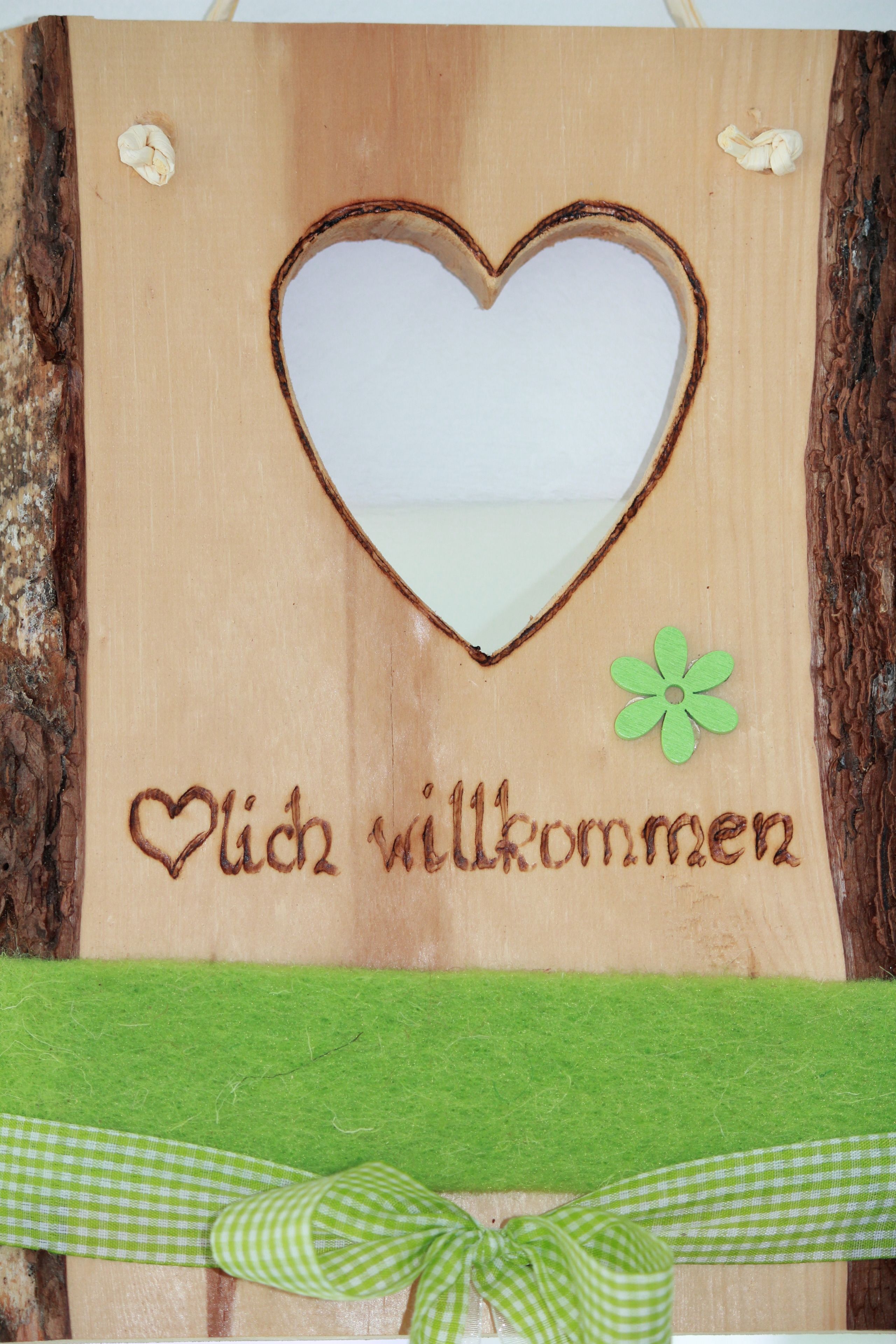 A wooden sign with a heart-shaped cutout, a green ribbon, and the text "Ich wilkommen" (Welcome).