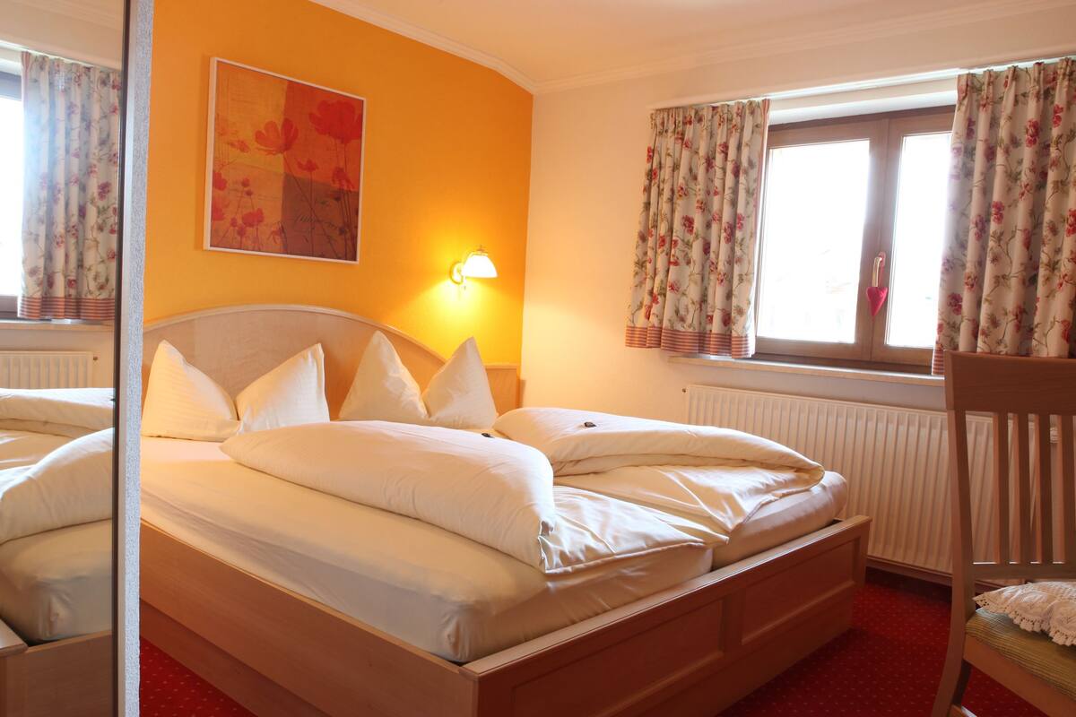 A double bed with white linens, a wooden headboard, a wall-mounted lamp, and a floral painting.