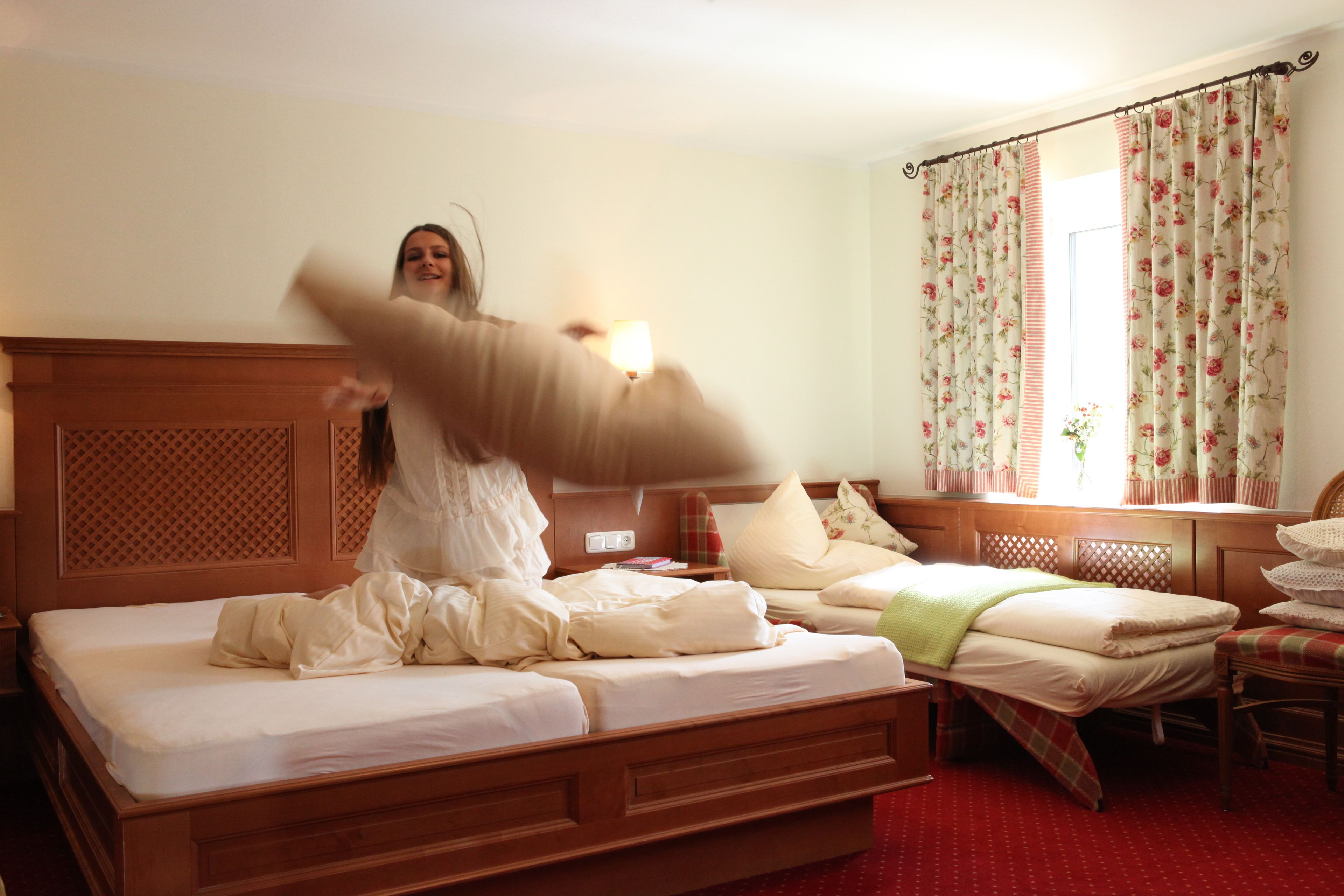 Habitación de hotel con una cama de madera, una mesita de noche, una lámpara, una ventana con cortinas florales y una silla.