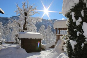 Un espace extérieur enneigé, doté d’une structure en bois, peut-être une terrasse ou un porche, entouré d’arbres chargés de neige et avec une chaîne de montagnes en arrière-plan.