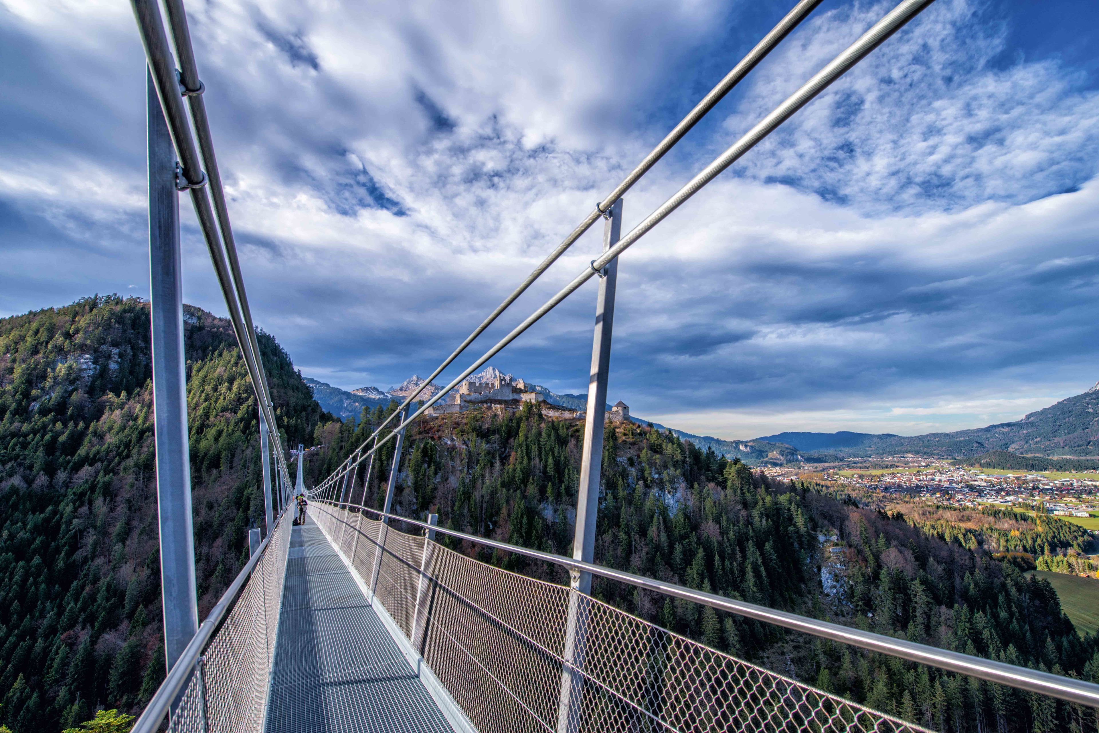 A suspension bridge with metal railings and a mesh walkway, overlooking a valley with a town and mountains in the background.