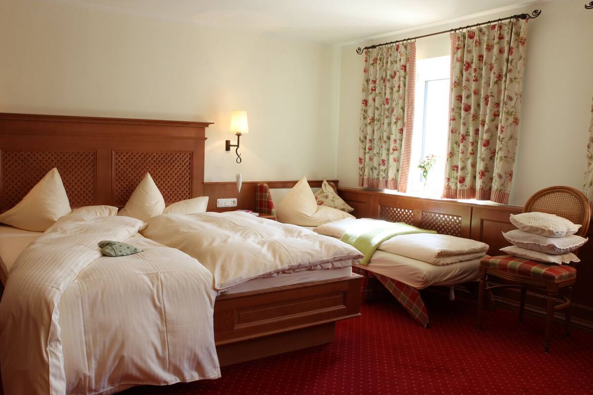 A double bed with a wooden headboard, two bedside tables with lamps, a chair with a cushion, and a window with floral curtains.
