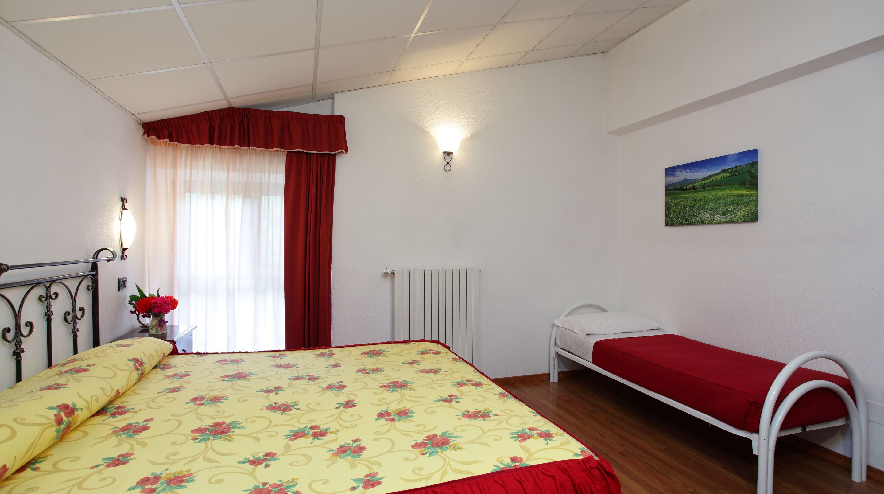 A bedroom with a bed, a bedside table, a radiator, a window with curtains, and a wall-mounted picture.