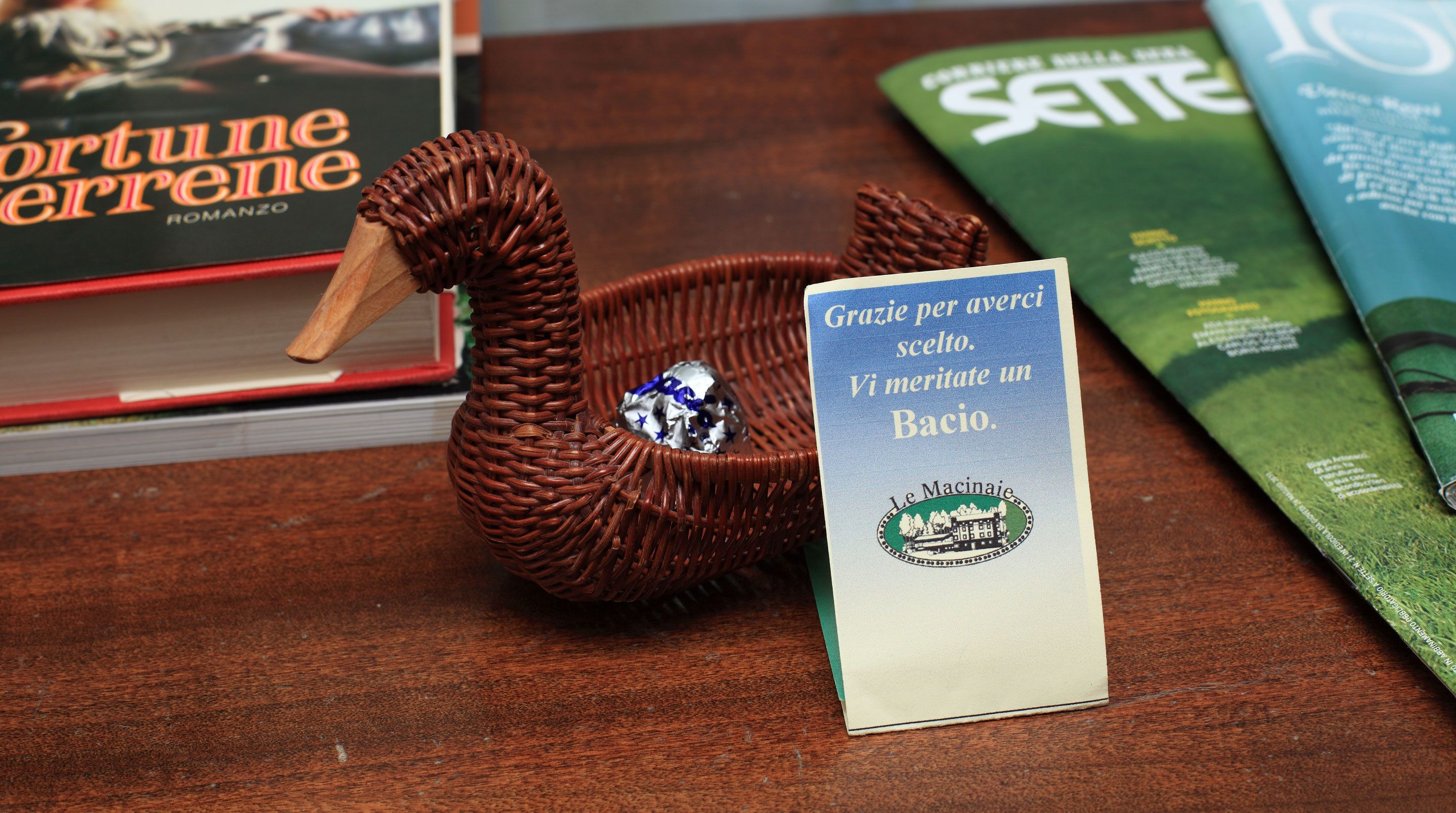 Ein schwanenförmiger Korb aus Weidwerk, der einen silbernen Stern enthält, platziert auf einer Holztischfläche mit Büchern und einem Schild in italienischer Sprache.