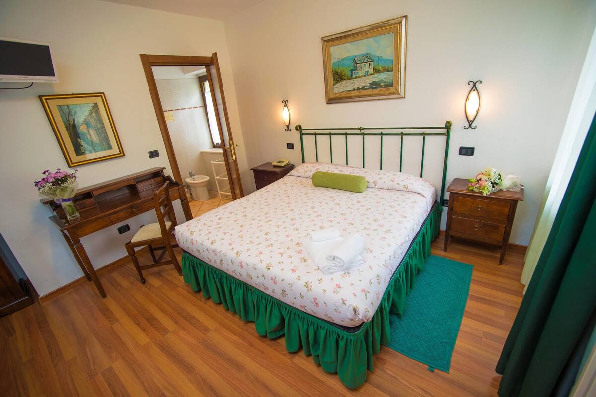 A bedroom with a bed, a desk, a chair, a mirror, a nightstand, and a wall-mounted TV.