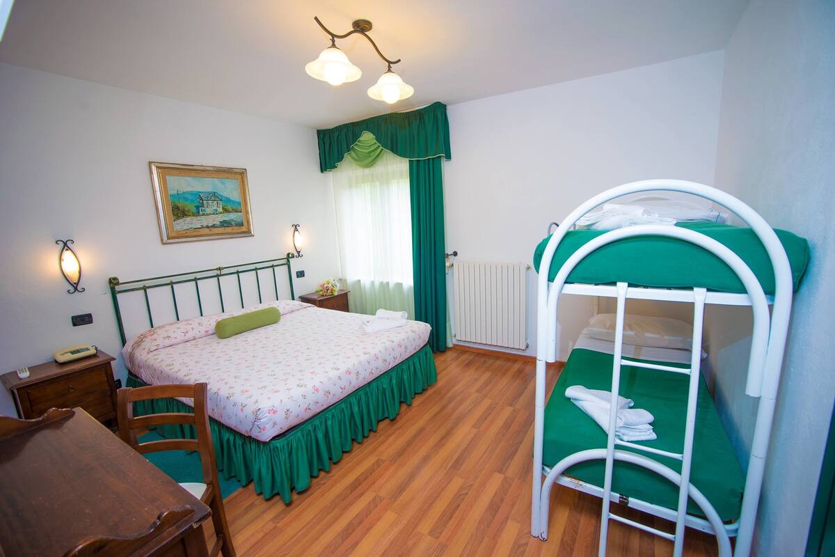 A bedroom with a bed, a desk, a chair, a radiator, a painting on the wall, and a window with curtains.