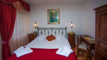 Un letto rifatto con lenzuola bianche e una coperta rossa, un comodino in legno con una lampada, una scrivania con una sedia e un quadro appeso al muro.