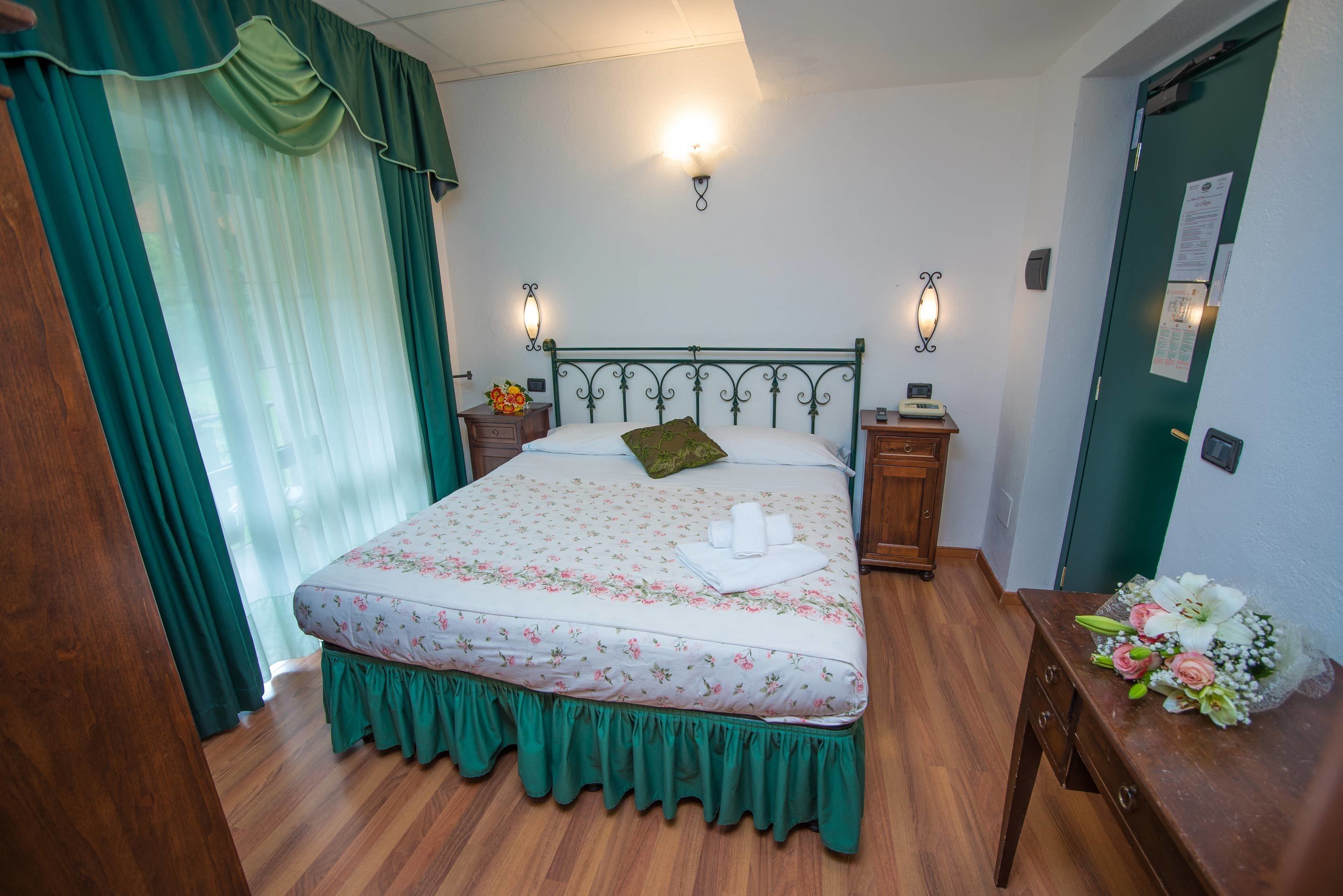 A neatly made bed with floral bedding, a green skirt, and a green pillow. There are two bedside tables with lamps, a wooden nightstand, and a wooden dresser.
