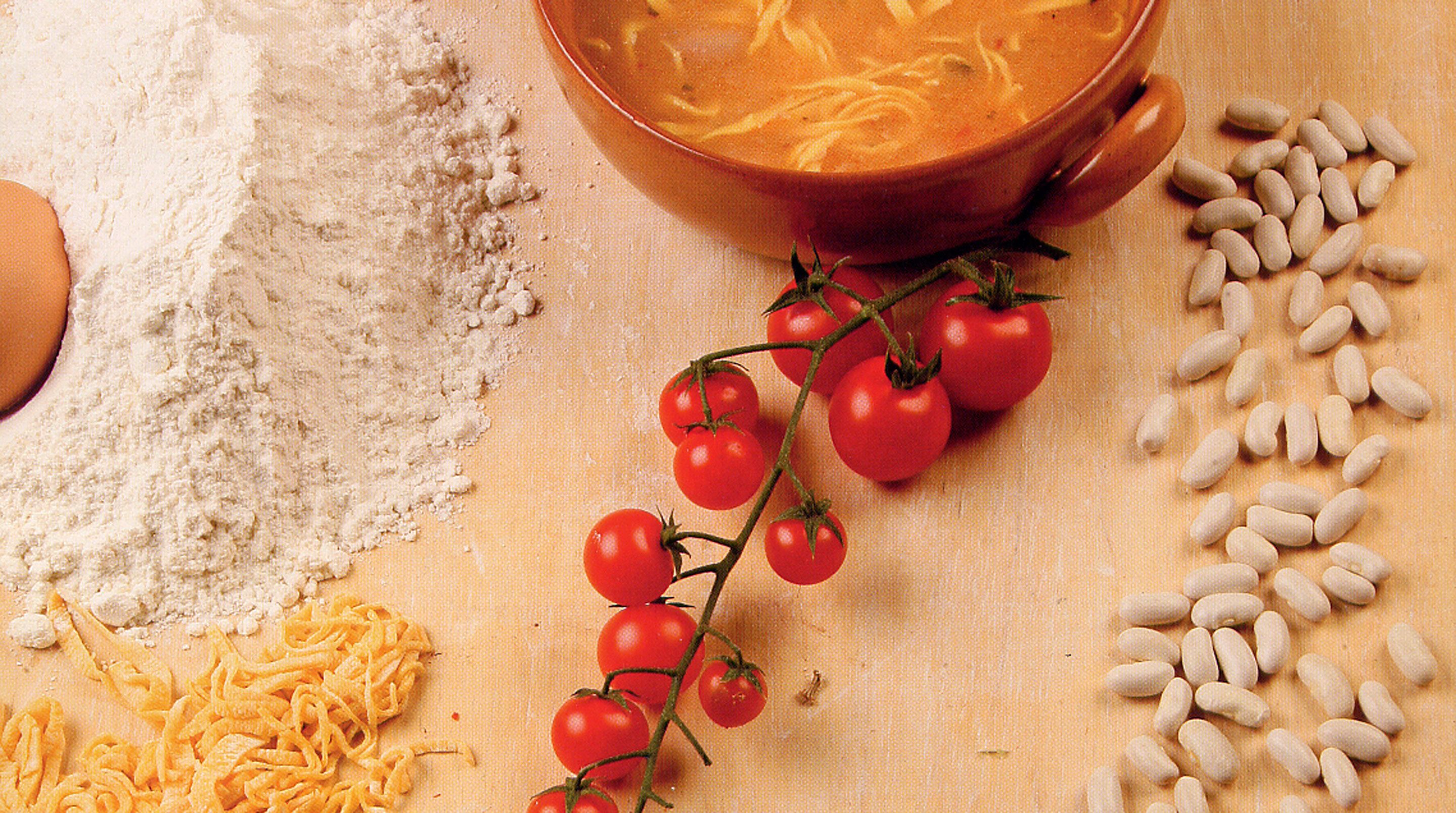 A table with a bowl of soup, a tomato plant, flour, and pasta.