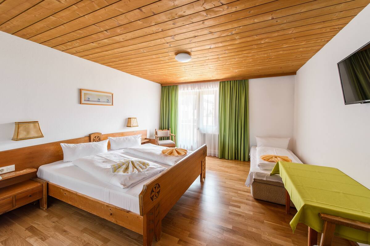 Quarto com teto de madeira, uma cama grande, uma cama menor, uma escrivaninha com toalha de mesa verde e uma televisão na parede.