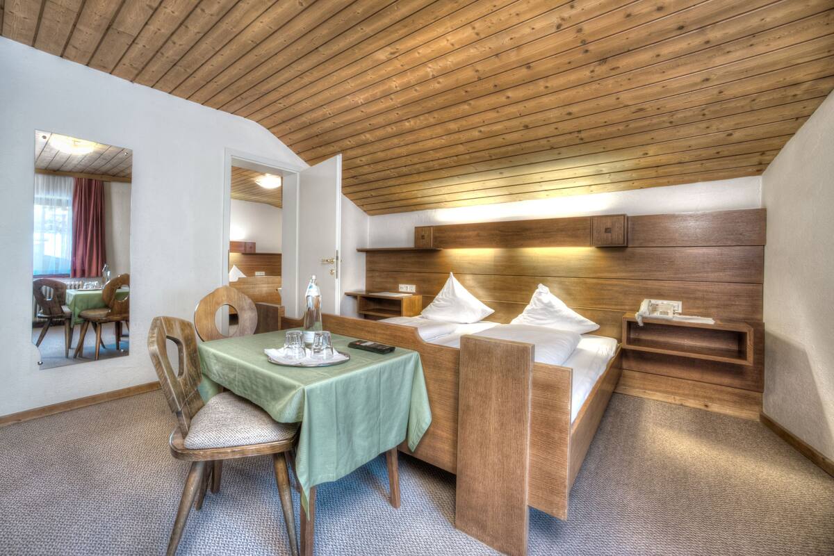 Quarto compacto com teto de madeira, cama com almofadas, mesa de cabeceira, pequena mesa com toalha de mesa verde e uma cadeira.