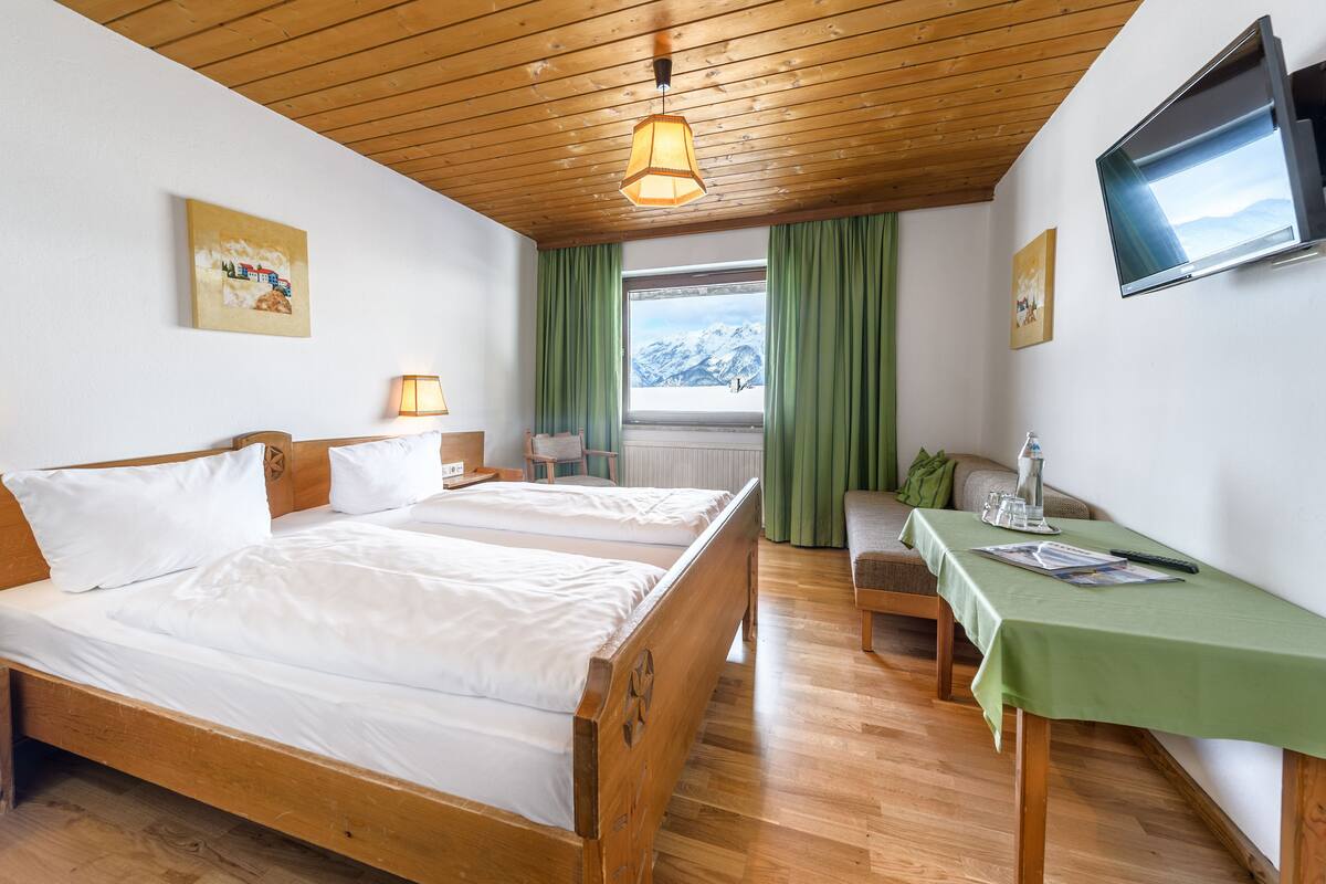 Quarto de hotel com duas camas de madeira, uma escrivaninha com toalha de mesa verde, uma TV fixada na parede e uma janela com vista para montanhas nevadas.