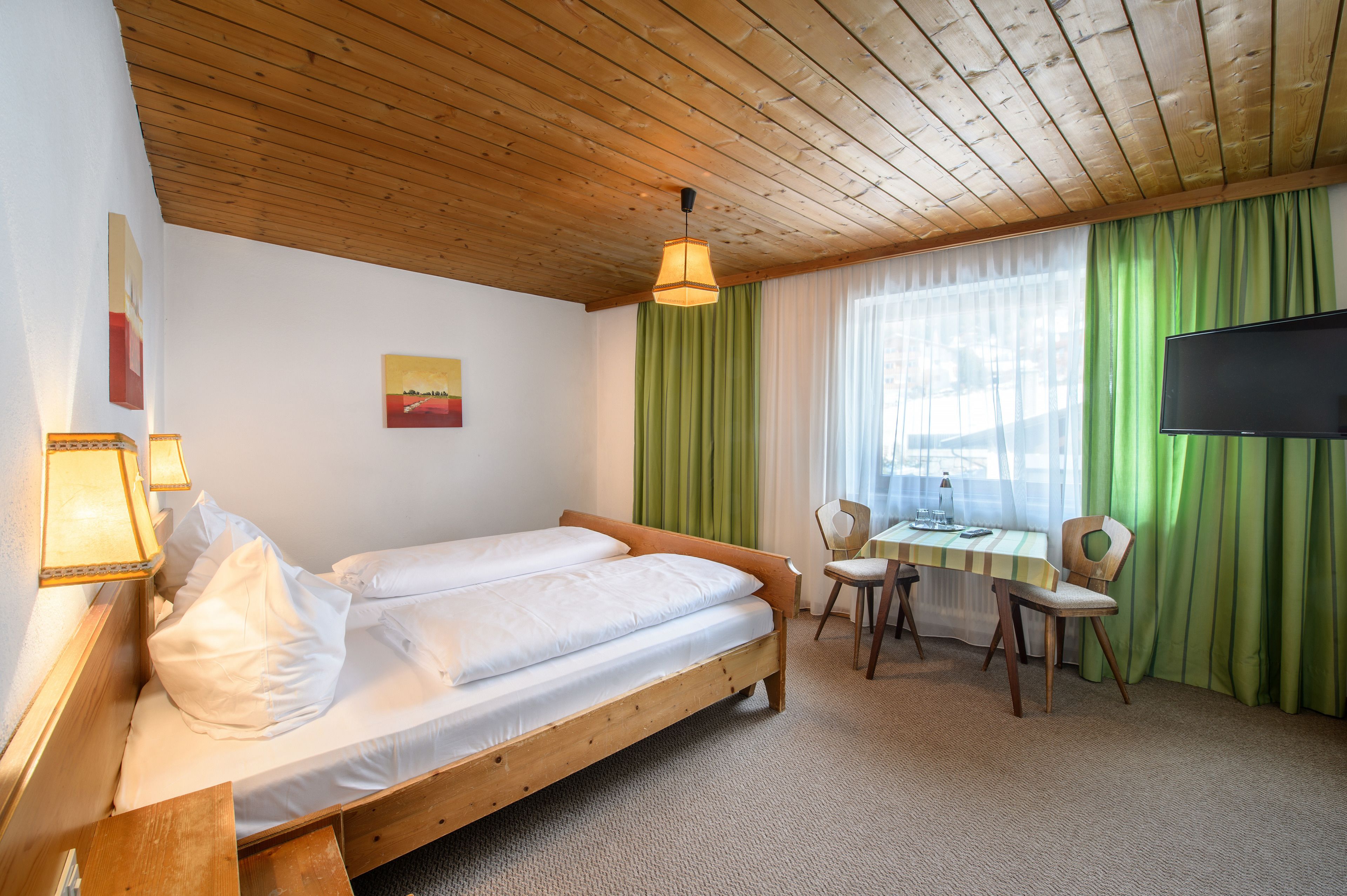 Quarto com cama de madeira, escrivaninha com cadeiras, televisão e janela com cortinas verdes.