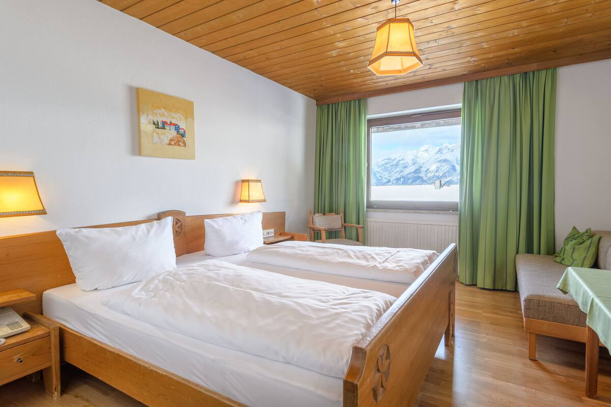 Quarto de hotel com duas camas, cabeceiras de madeira, teto de madeira, janela com vista para montanhas e cortina verde.