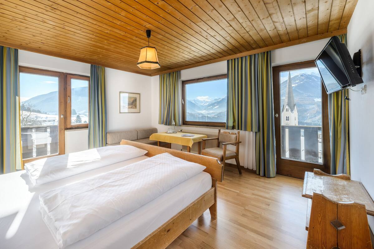 Quarto de hotel com uma cama grande, uma escrivaninha e uma cadeira. Há janelas com cortinas, teto de madeira e uma televisão na parede.