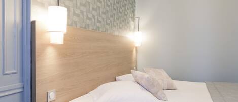 Una cama bien tendida con sábanas blancas y dos almohadas, un cabecero de madera y dos lámparas fijadas a la pared.