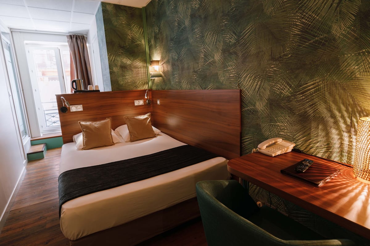 A hotel room with a wooden headboard, a bed with beige bedding, a bedside table with a lamp, a phone, and a green chair.