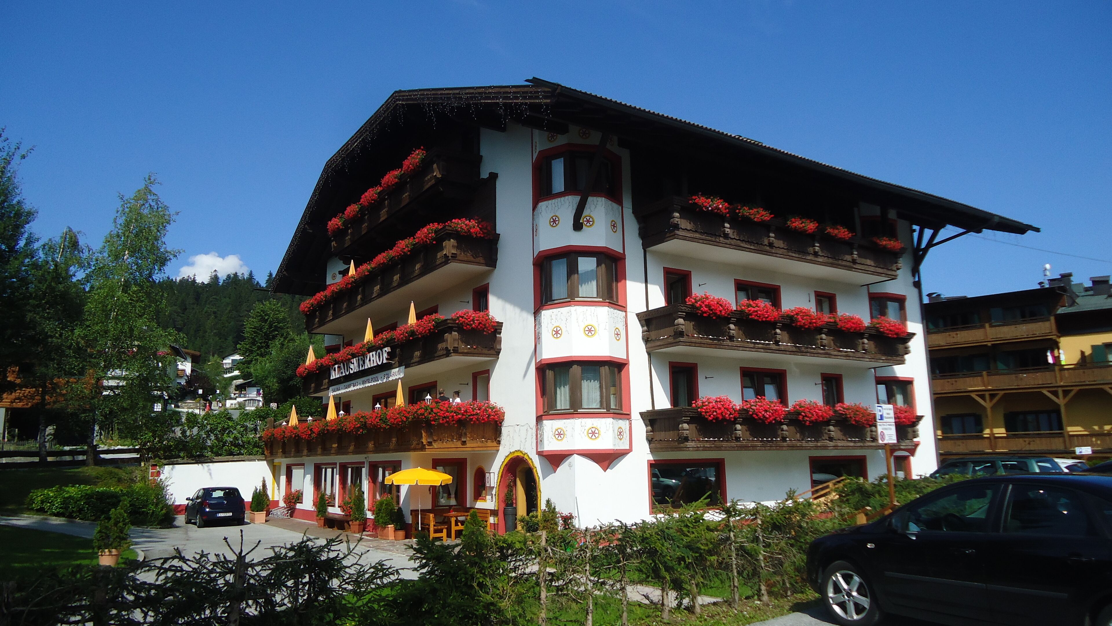 A multi-story hotel with balconies adorned with red flowers, a central entrance with a yellow umbrella, and a clear blue sky.