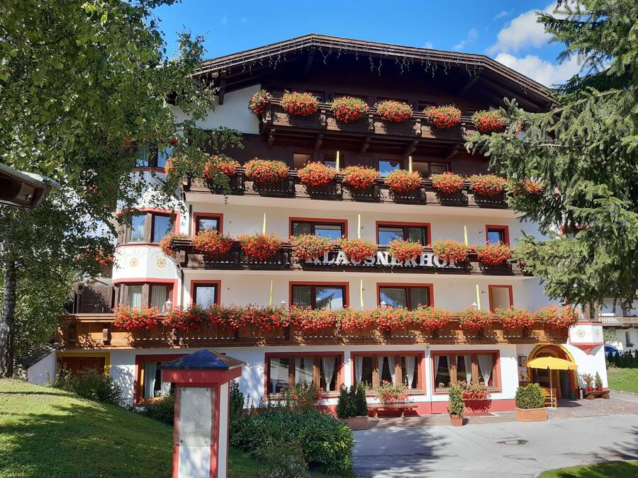 A multi-story hotel with balconies adorned with red flowers, a sign reading "LAUSNERHOT," and a well-maintained garden area.