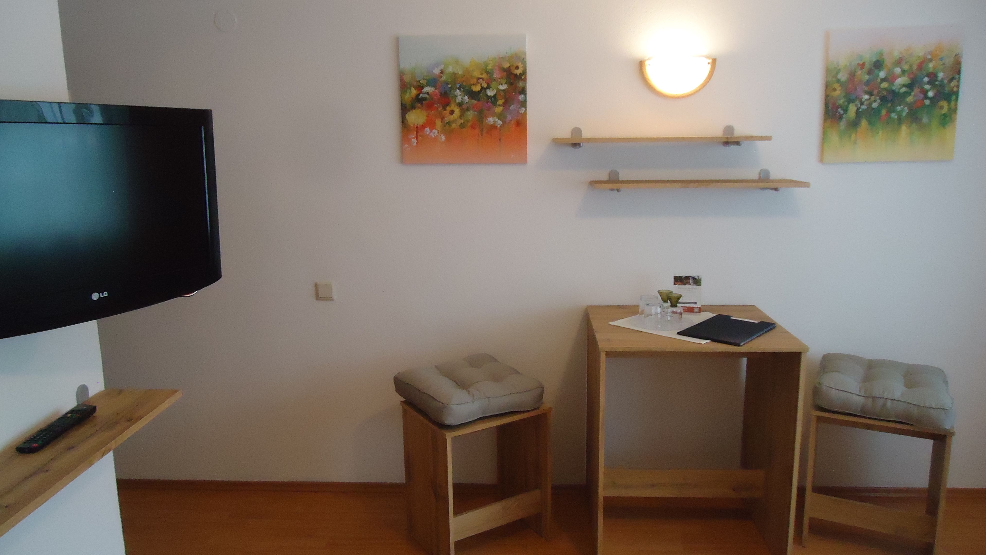 A small, tidy room with a flat-screen TV, two wooden chairs, a small table with a notebook, and two wall-mounted shelves with paintings.
